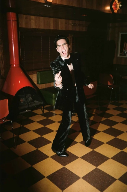 A man in a sequined suit stands on a checkerboard floor with a vintage fireplace and wood paneling, sticking out his tongue and gesturing.
