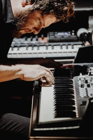 Man playing a black and white keyboard in a dark room, close up, with his hands and curly hair visible in the close shot.