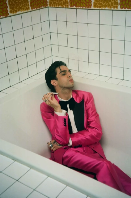 Young adult man in pink suit poses in a white tiled bathroom, holding a cigarette, creating an edgy yet stylish aesthetic.