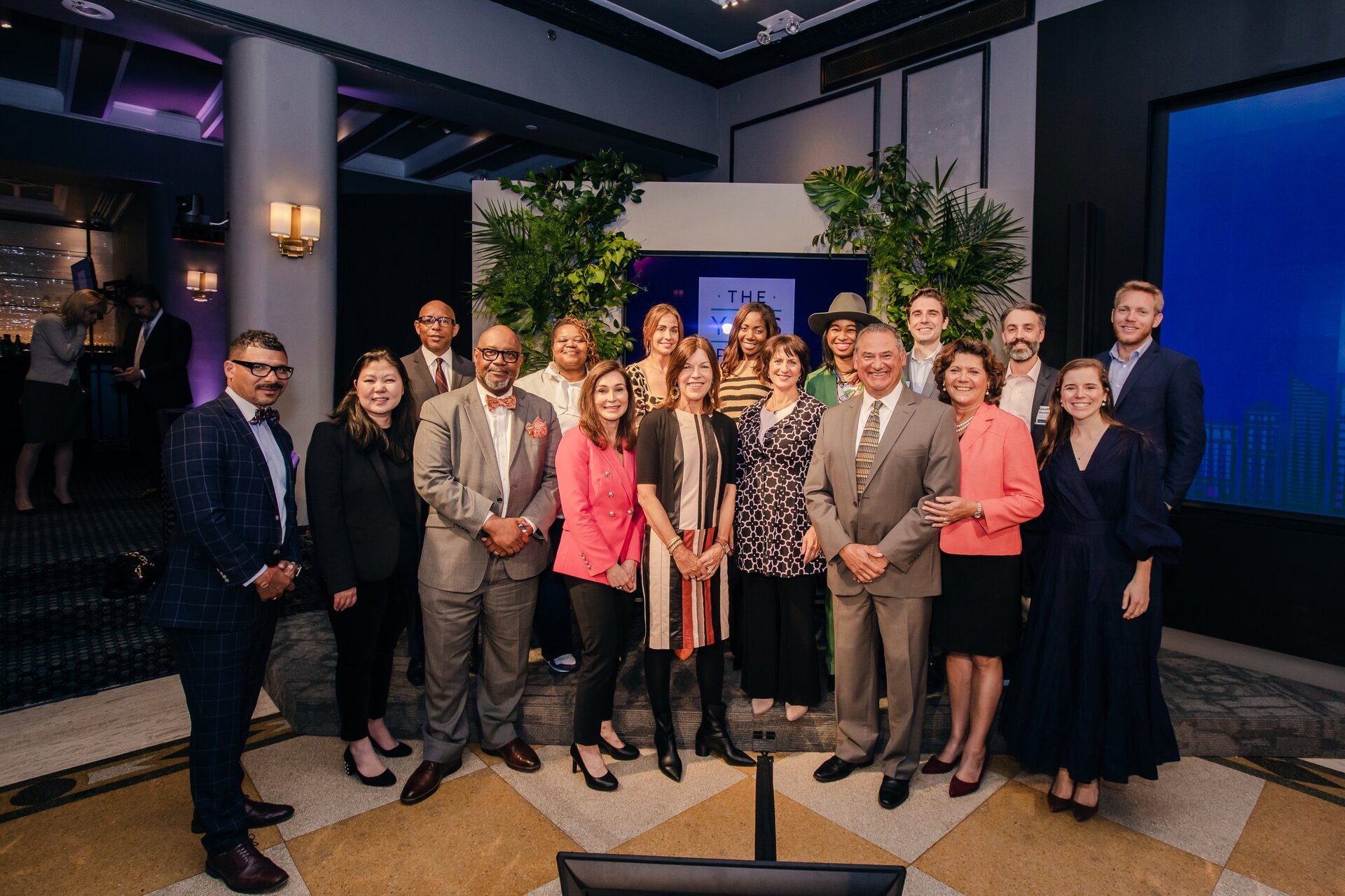 Indoor event photograph featuring a group of people of diverse ages and ethnicities posing together in a warmly lit interior setting.
