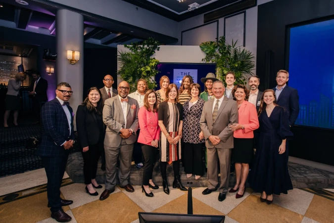 Indoor event photograph featuring a group of people of diverse ages and ethnicities posing together in a warmly lit interior setting.