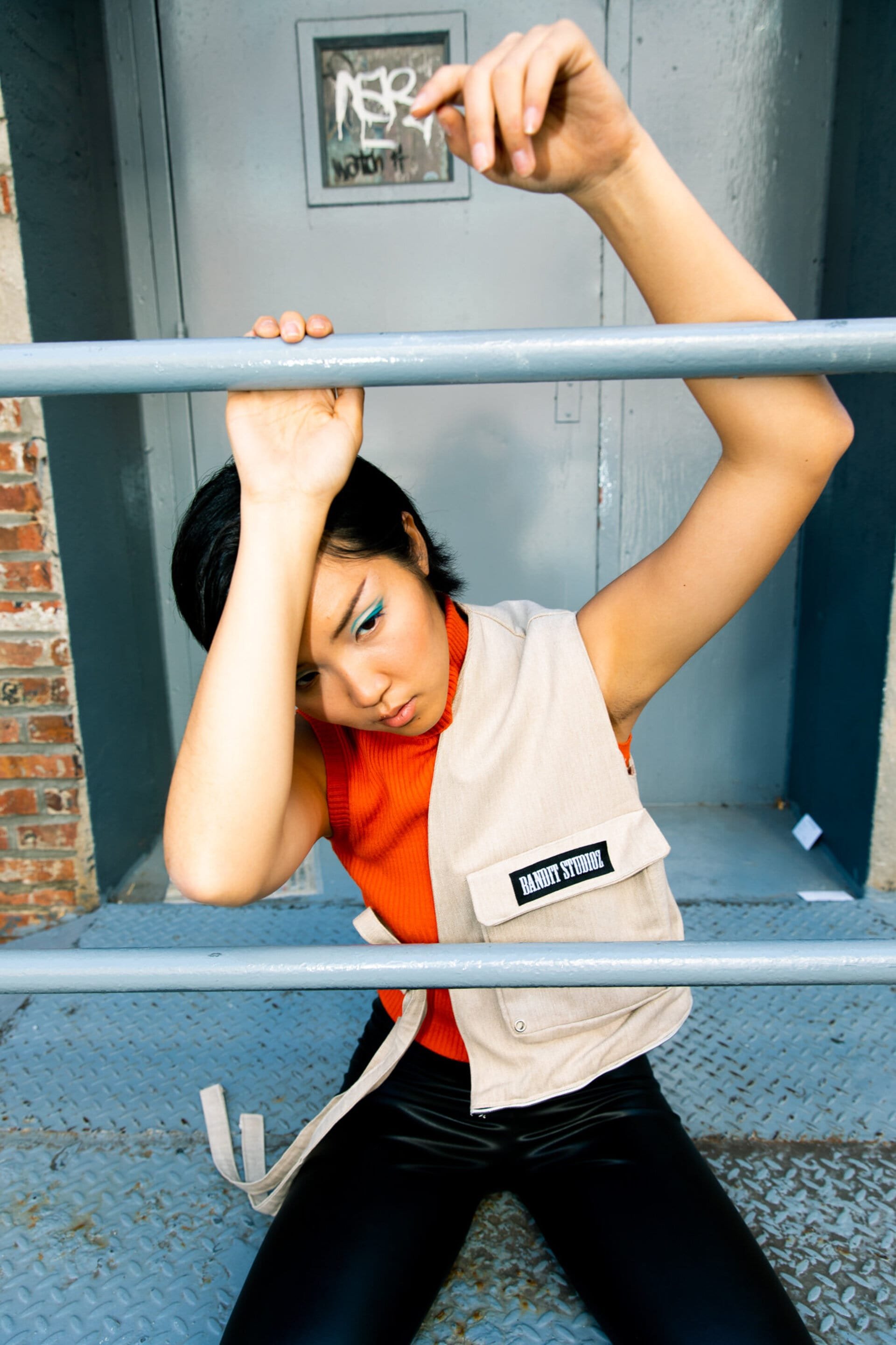 Young Asian woman in vest poses behind metal bars in urban setting, wearing bright makeup, with textured background and natural lighting.
