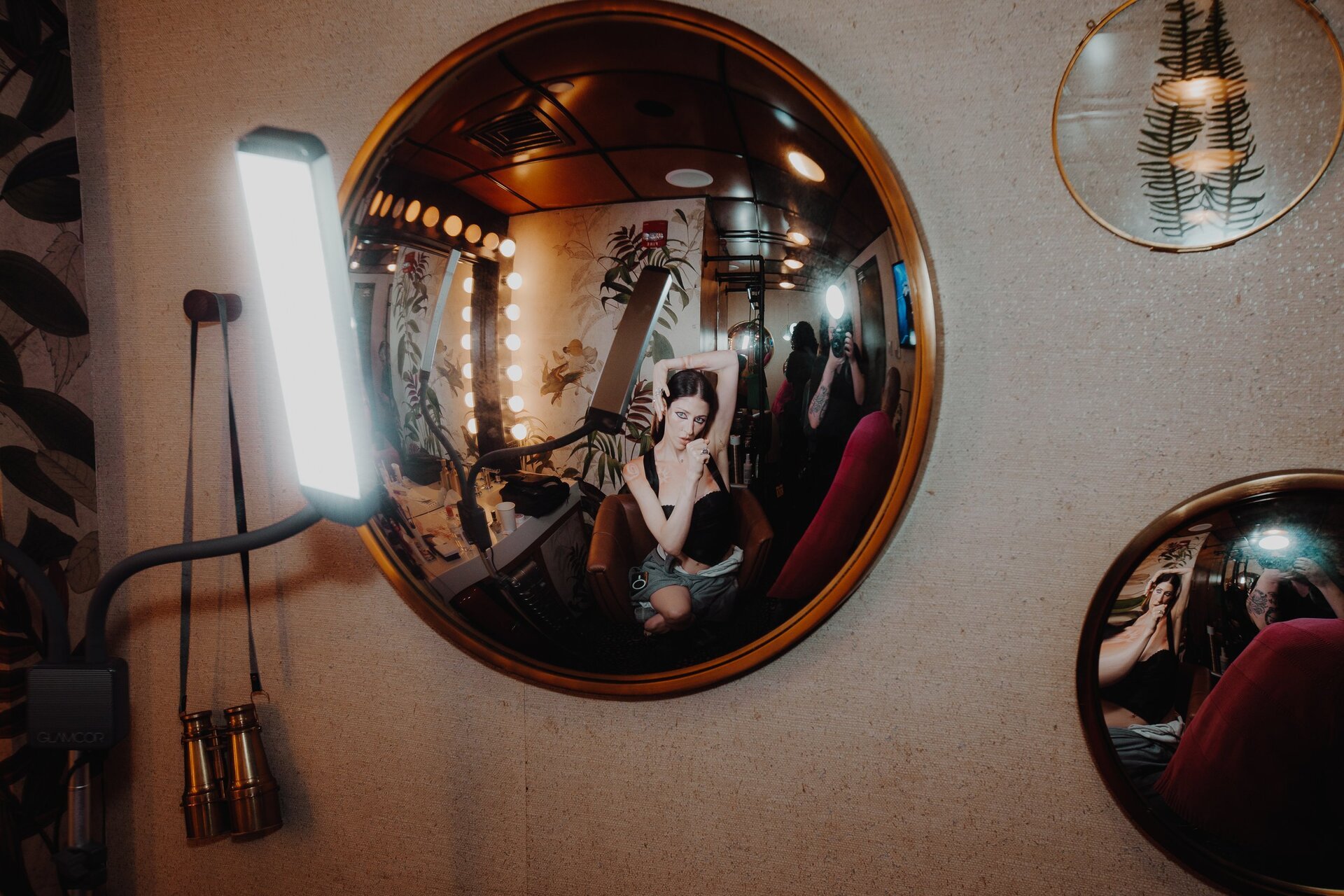Woman poses reflected in a round mirror in a vintage-style room with a lamp, floral wallpaper, and other round mirrors in the background.