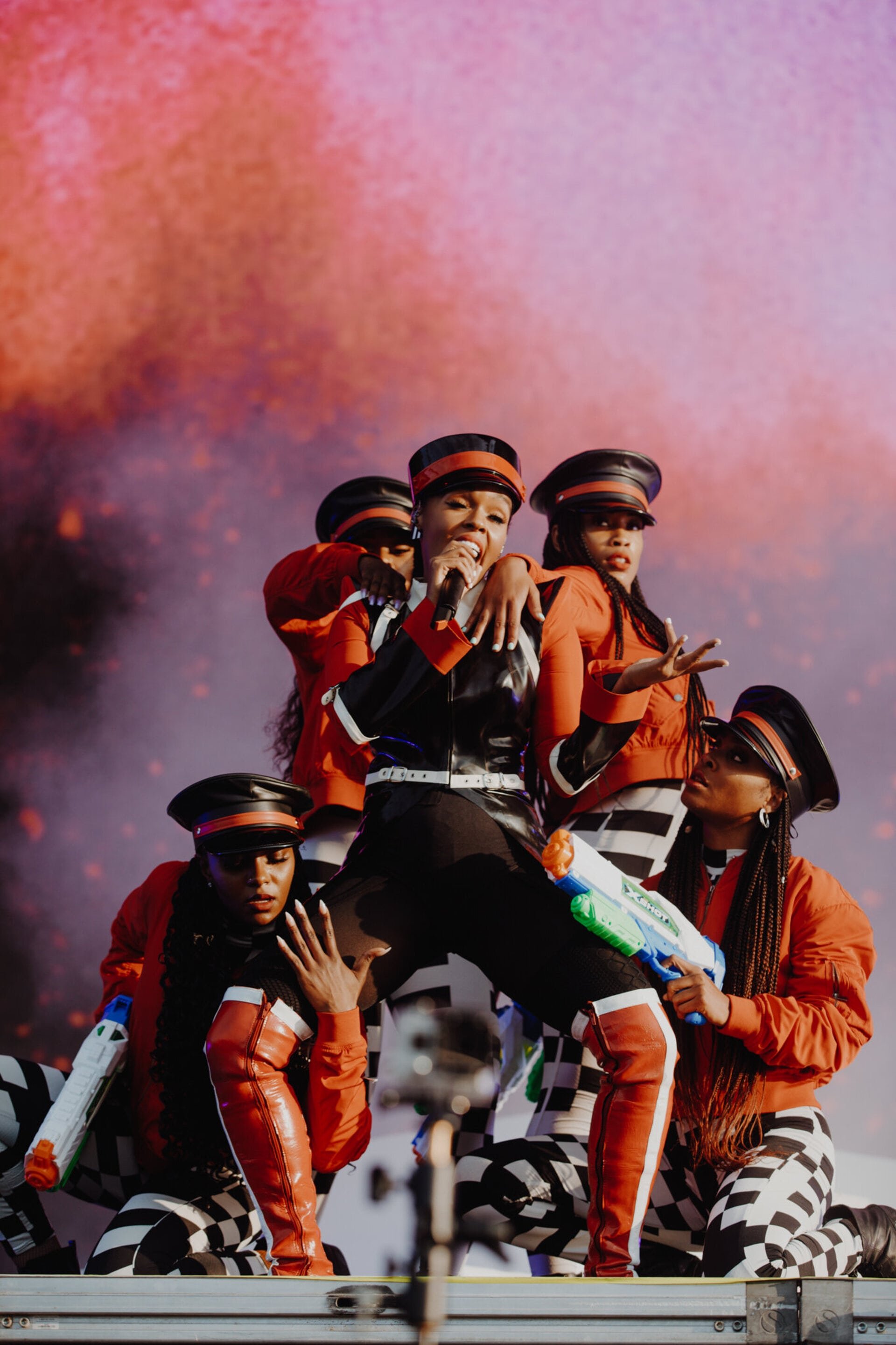 Janelle Monáe performs on stage with backup dancers holding toy water guns in red, black and white costumes in a concert setting.