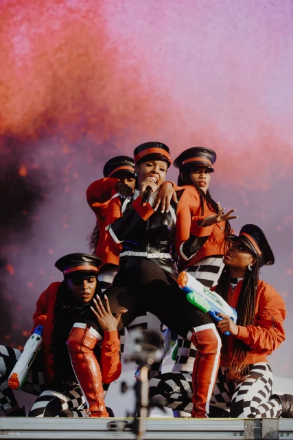 Janelle Monáe performs on stage with backup dancers holding toy water guns in red, black and white costumes in a concert setting.