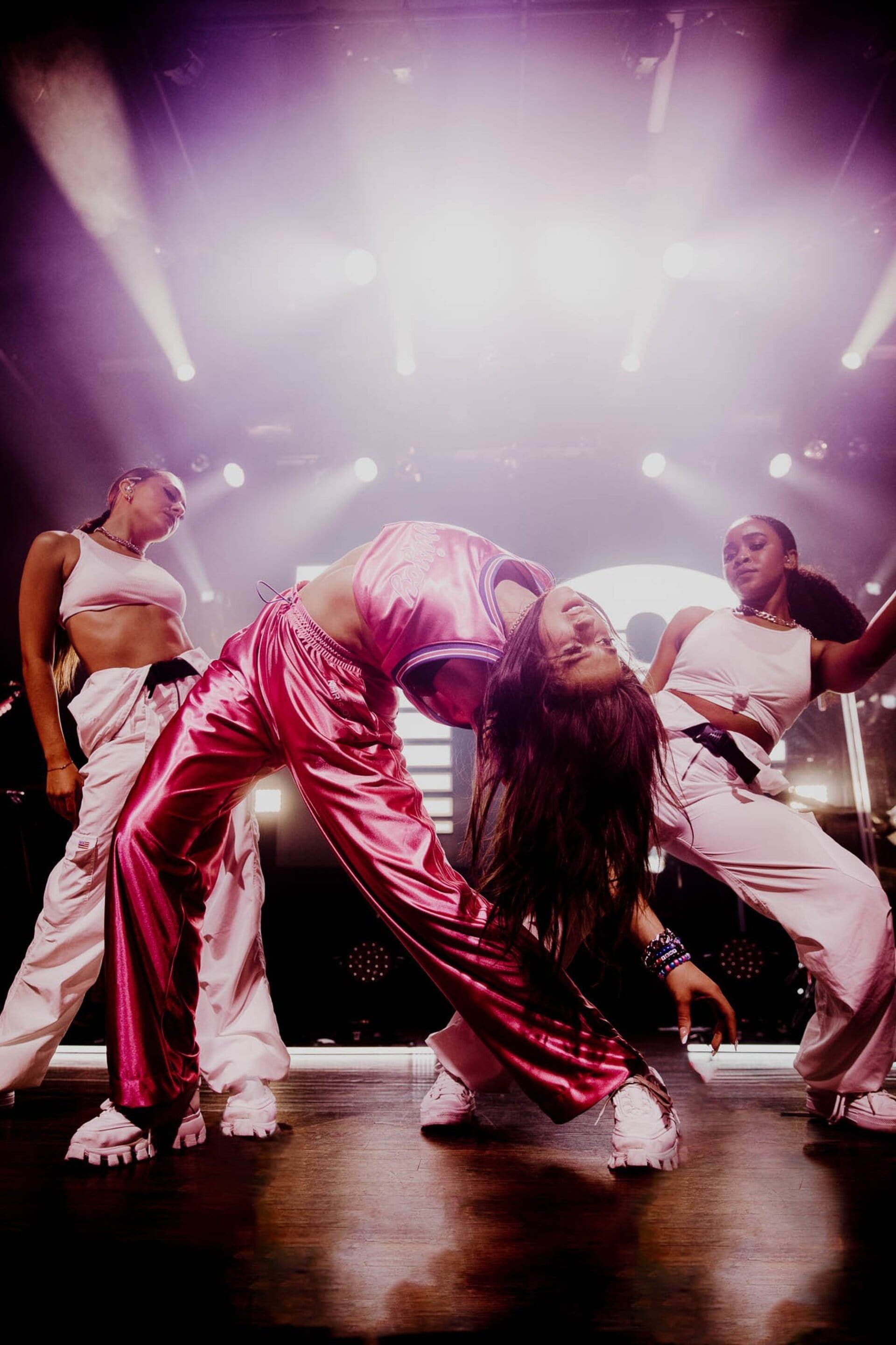 Three women perform a dance routine on a stage, dressed in athletic wear with spotlights illuminating them from above.