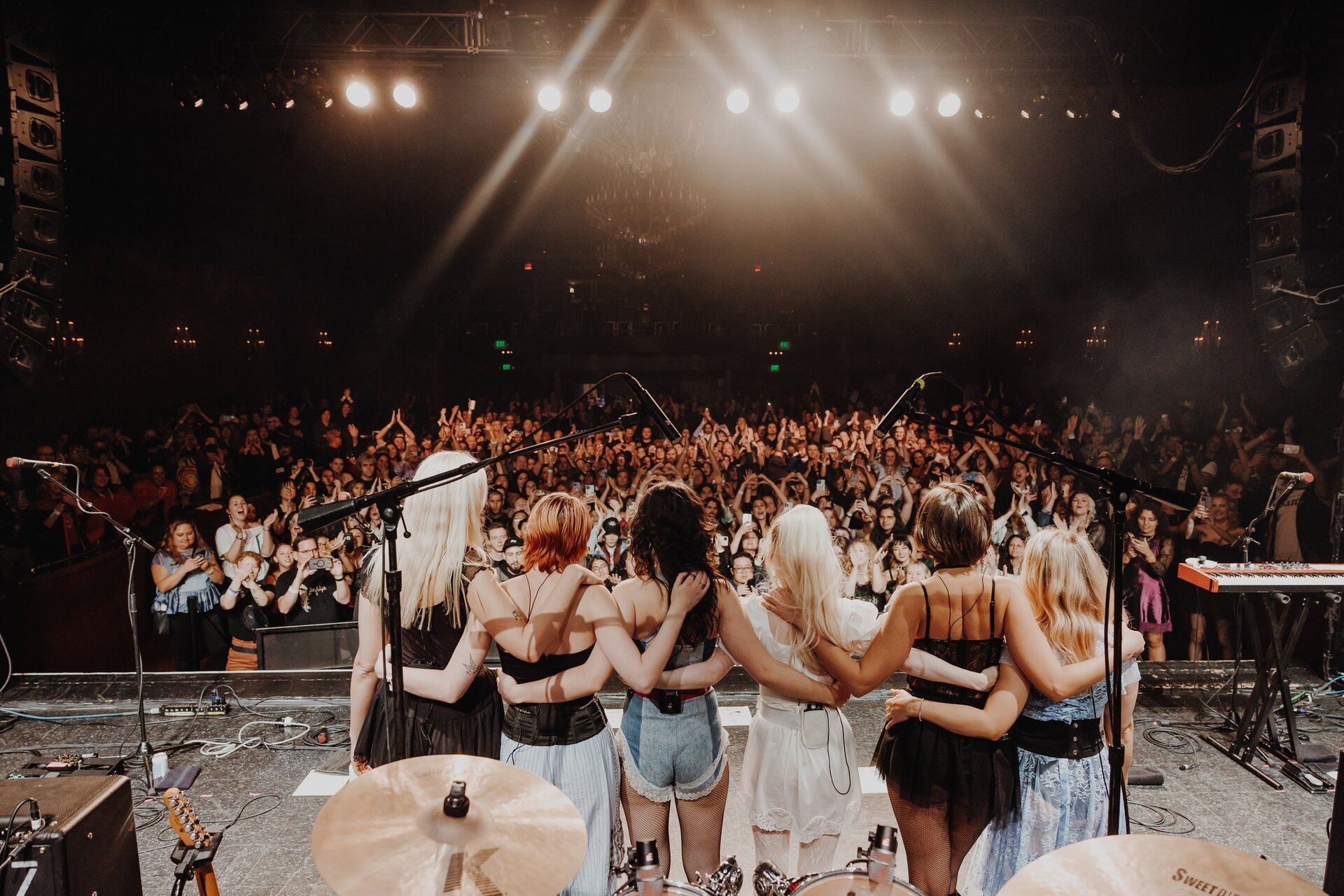 A band of six women stands on stage, arms linked, facing a large crowd under bright stage lights in a concert venue.