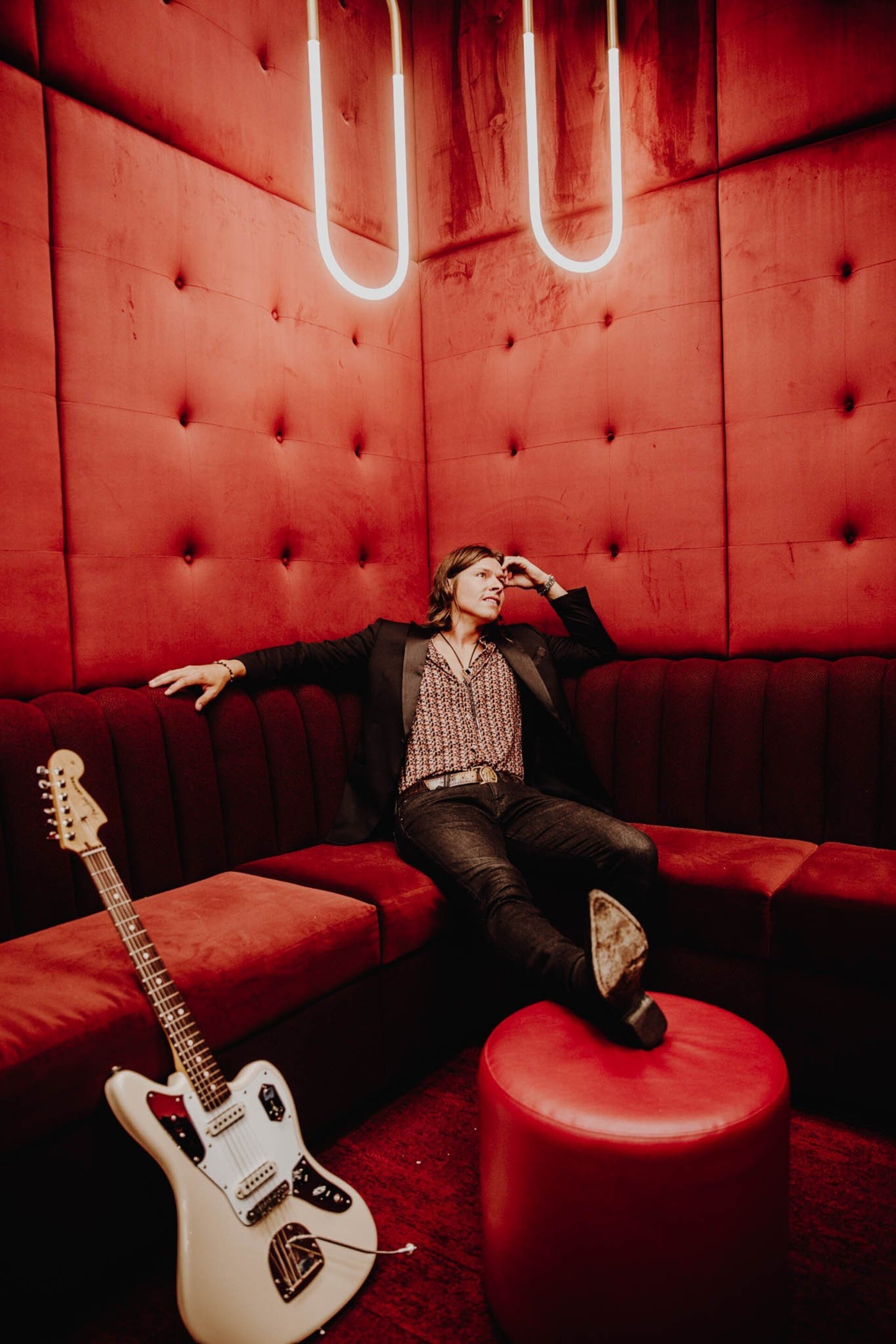Person poses on a tufted red velvet couch with a guitar nearby, illuminated by red light in a room with a neon light fixture.