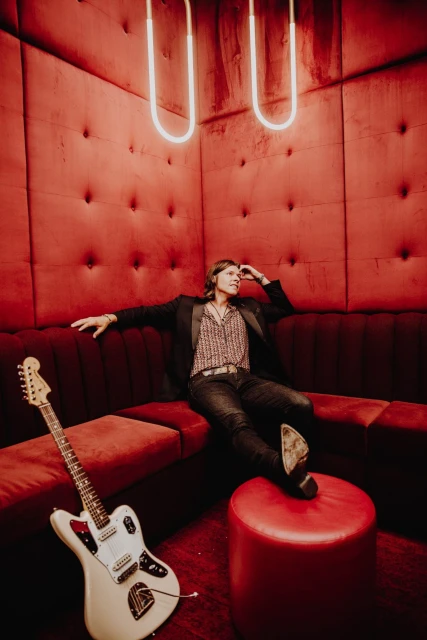Person poses on a tufted red velvet couch with a guitar nearby, illuminated by red light in a room with a neon light fixture.