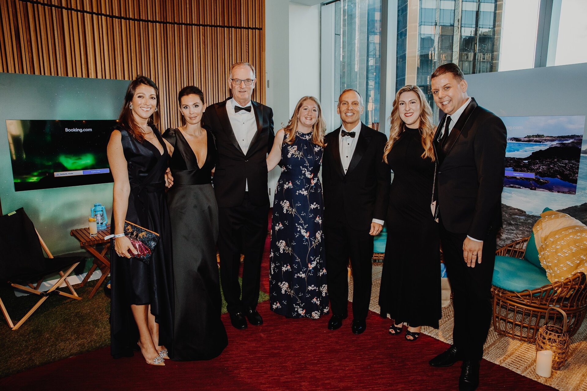 Group of six adults in formal wear pose indoors on a red carpet in front of two monitors, with an urban background visible.