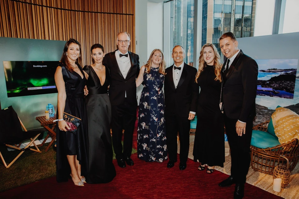 Group of six adults in formal wear pose indoors on a red carpet in front of two monitors, with an urban background visible.