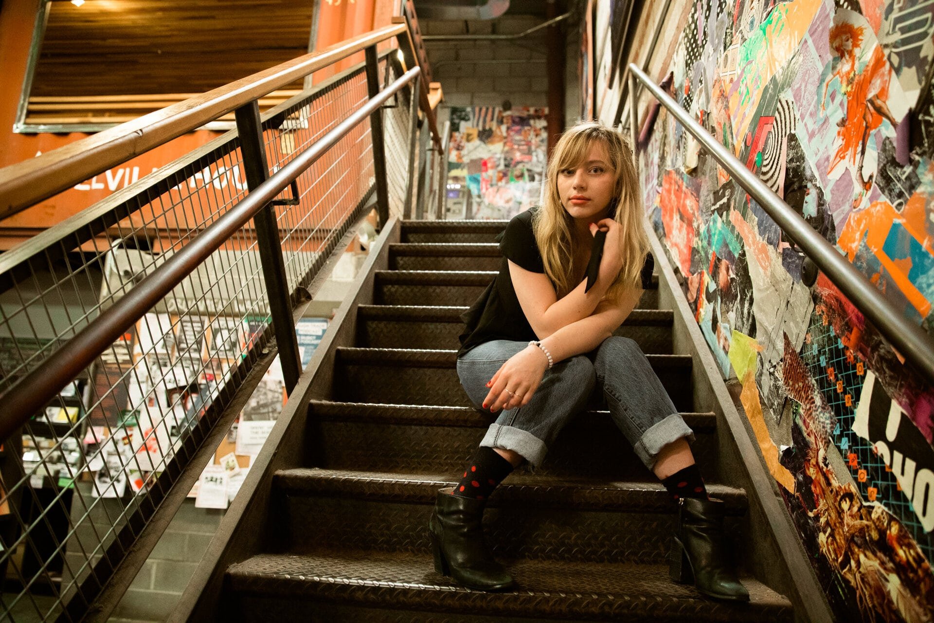 Young woman with blonde hair poses while sitting on metal stairs, inside a building with a collage wall and metal handrails.