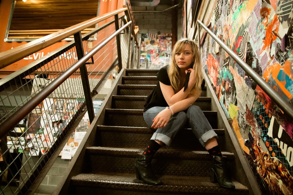 Young woman with blonde hair poses while sitting on metal stairs, inside a building with a collage wall and metal handrails.