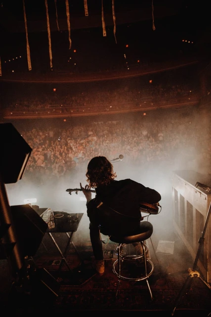 Guitarist performs on stage to a large crowd in a concert hall with stage fog and dramatic lighting, creating an atmospheric scene.