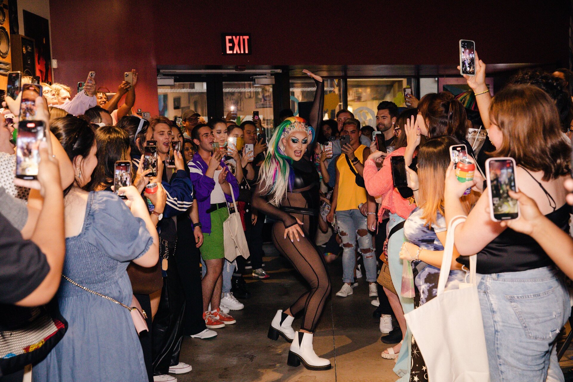 Drag queen performs in front of an audience holding cell phones, indoor venue, full frame shot, rainbow wig, black leotard, white boots.
