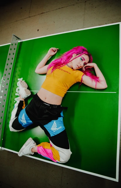 A young adult female with vibrant pink hair and colorful streetwear lies on a green ping pong table in an indoor setting with gray walls.