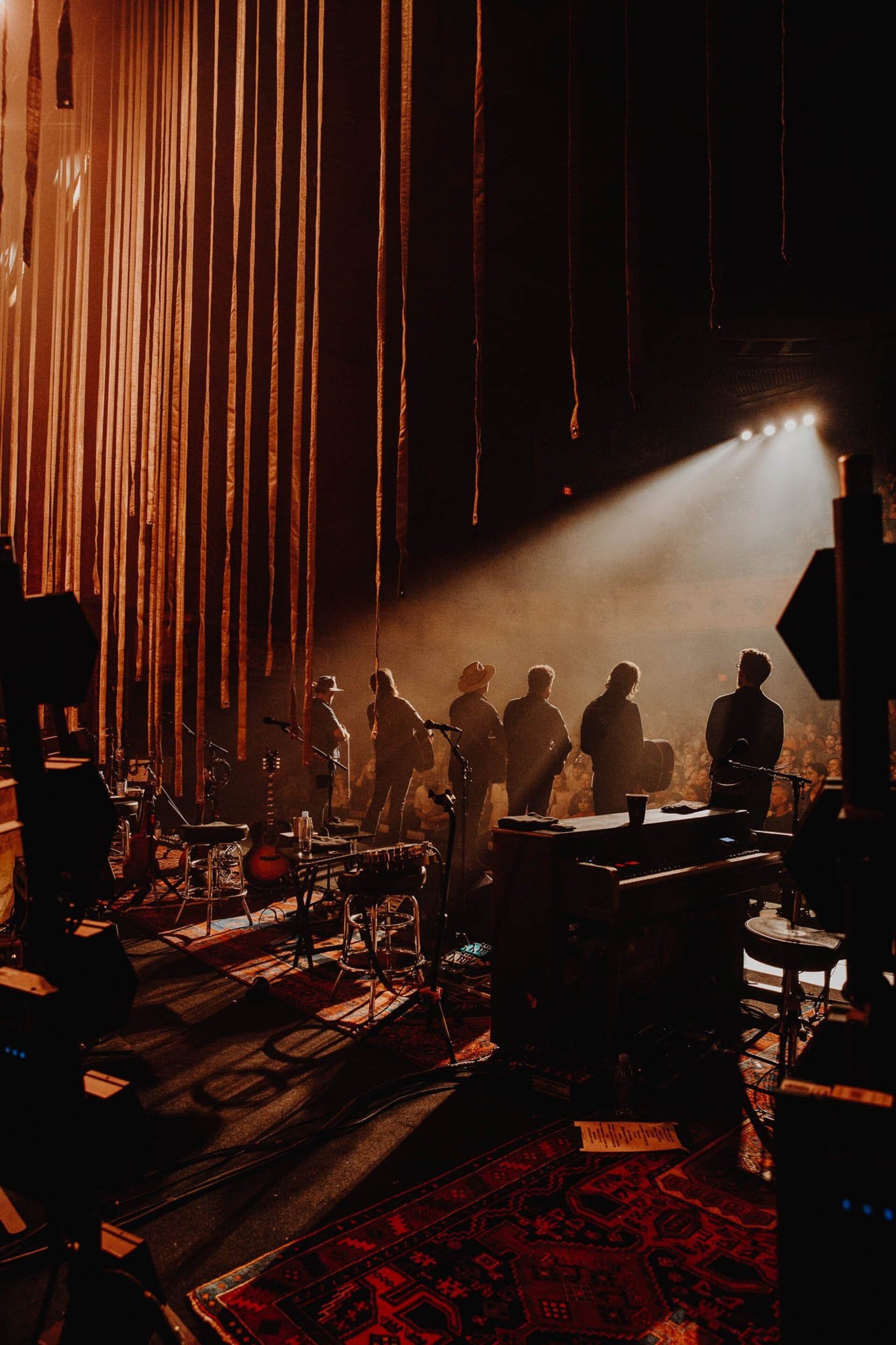 On a dimly lit stage, a band of musicians stands under a spotlight, surrounded by instruments, with long, vertical strips as a backdrop.