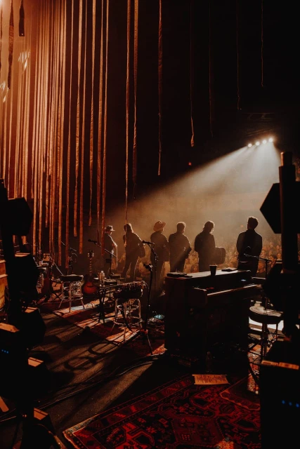 On a dimly lit stage, a band of musicians stands under a spotlight, surrounded by instruments, with long, vertical strips as a backdrop.