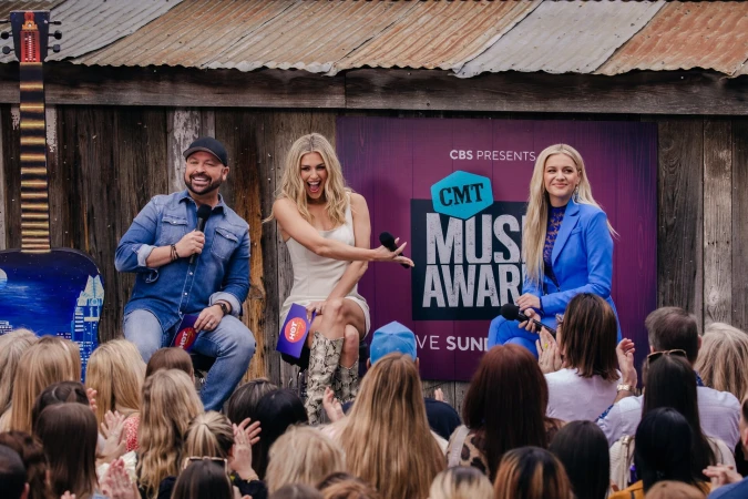 Three music artists at the CMT Music Awards speak to a crowd on an outdoor stage with a rustic wooden backdrop and sponsor signage.