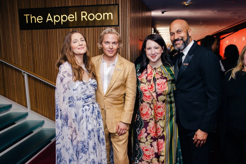 Group of four people at an event, posing in front of the Appel Room sign. Two women in floral dresses, and two men in suits.