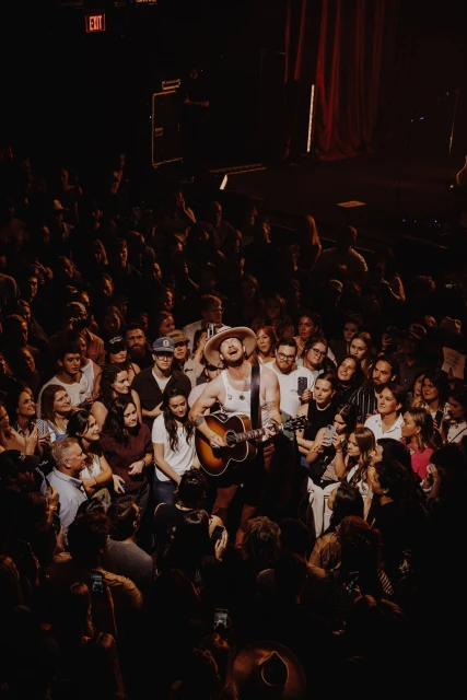 A musician plays guitar and sings into a microphone on stage at an indoor music venue, surrounded by an audience of fans in the dark.