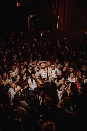 A musician plays guitar and sings into a microphone on stage at an indoor music venue, surrounded by an audience of fans in the dark.