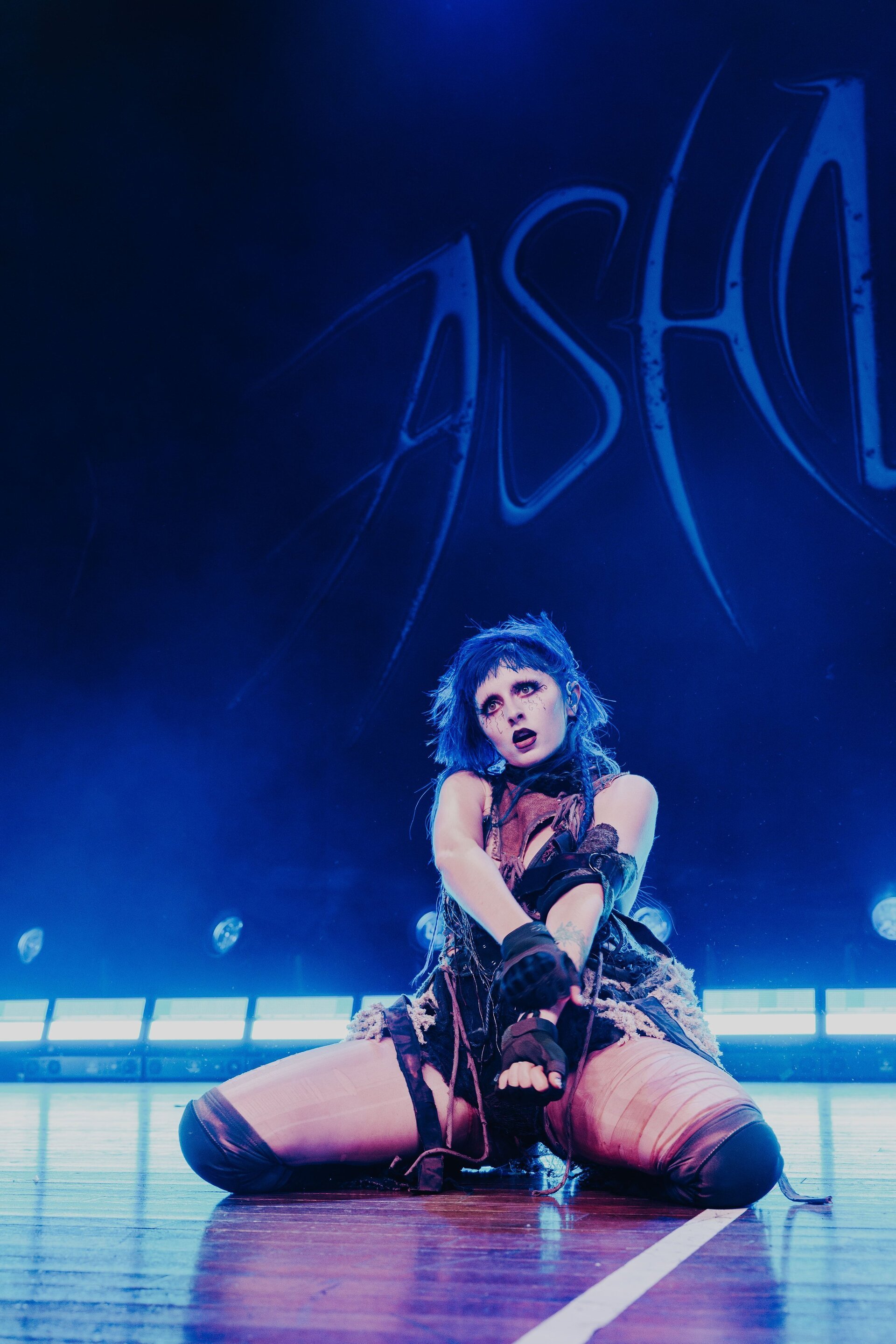 A singer with blue hair kneels on stage during a performance, wearing a unique outfit in vibrant blue stage lighting, in concert.