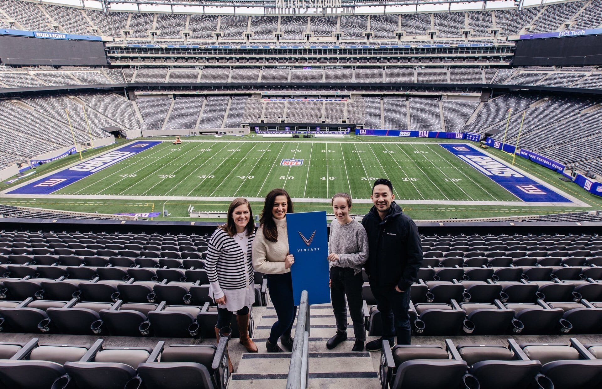 Group of four people stand in stadium seats holding a Vinfast sign, with the football field and stadium seating in the background.