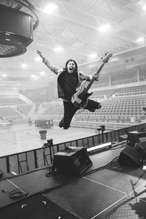 Musician with open mouth jumping mid-air on stage with a white bass guitar, lit by stage lights and in front of arena seating.