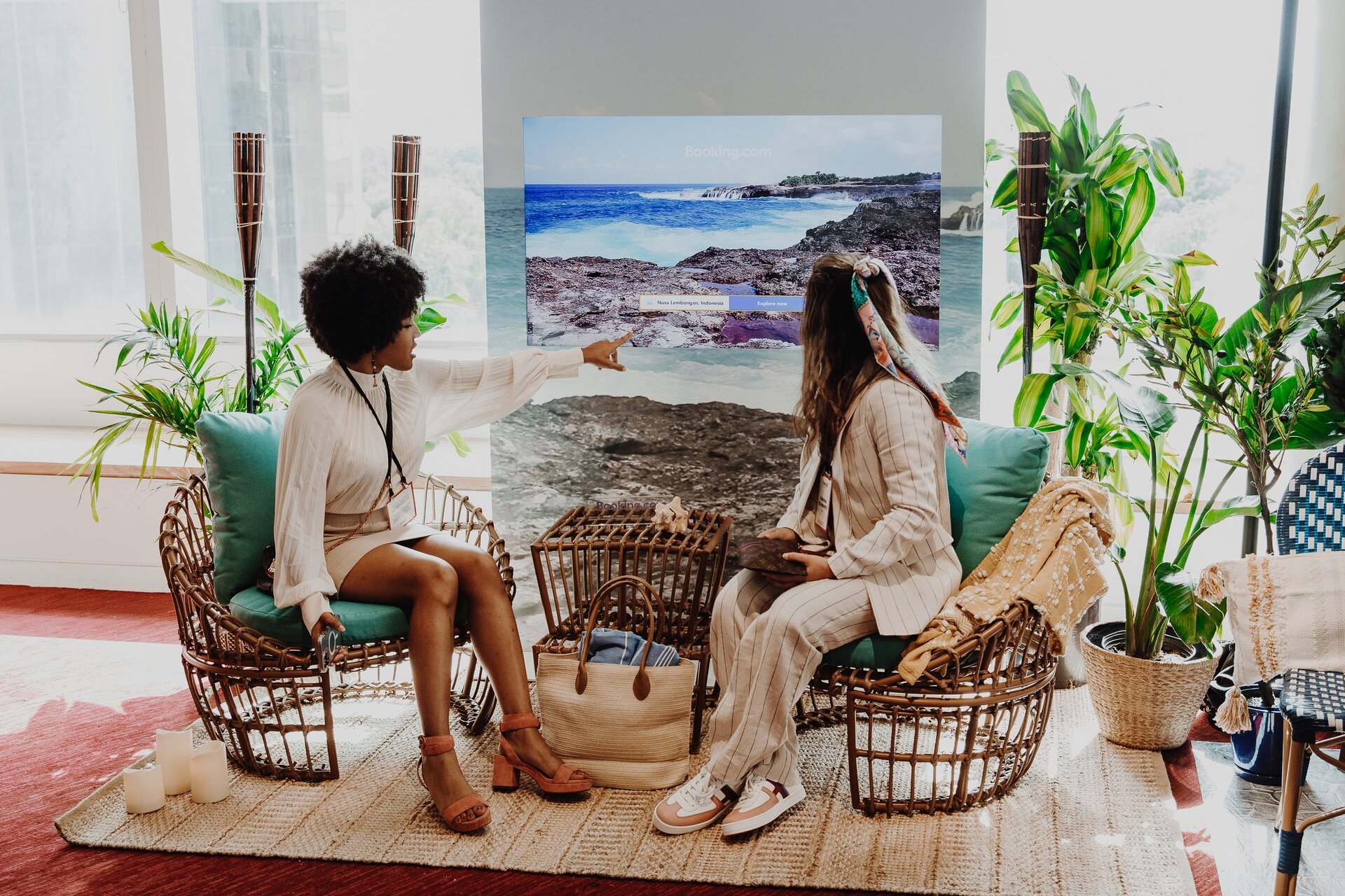 Two women sitting in wicker chairs in an indoor setting, with tropical plants and a beach scene on a screen in the background.