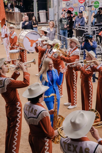 Blonde woman sings in a blue suit surrounded by the Texas Longhorn marching band in burnt orange and white uniforms outdoors.