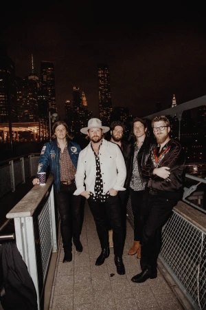 Band poses together in an urban setting at night with city buildings lit up in the background, wearing trendy casual clothing.