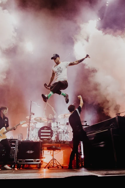 Band performing on stage with a singer leaping mid-air against a backdrop of smoke and bright lights during a live concert.