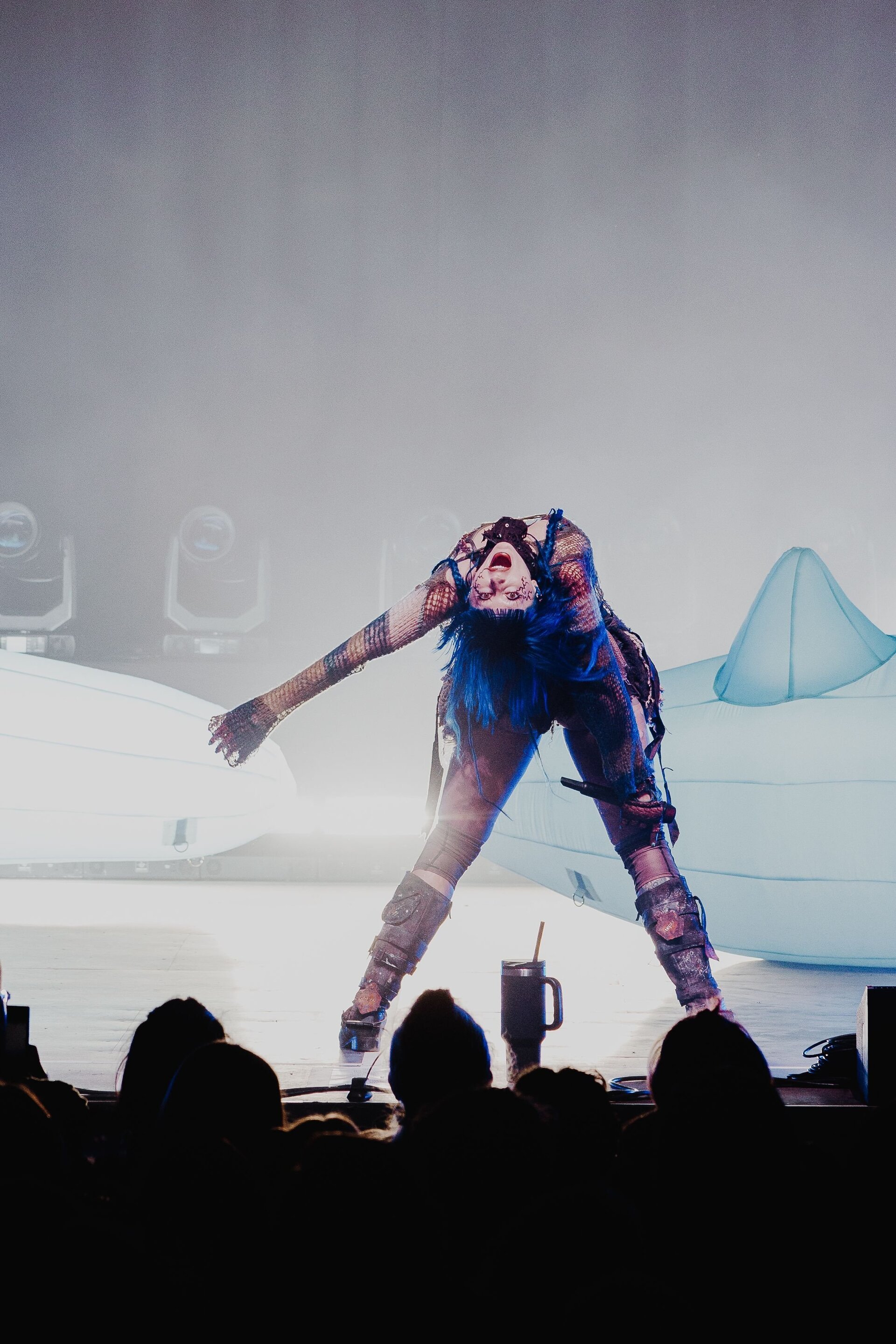 Performer with blue hair and dark makeup bends backward on a stage during a music concert with dark, silhouetted figures in foreground.