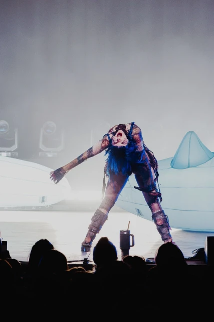 Performer with blue hair and dark makeup bends backward on a stage during a music concert with dark, silhouetted figures in foreground.