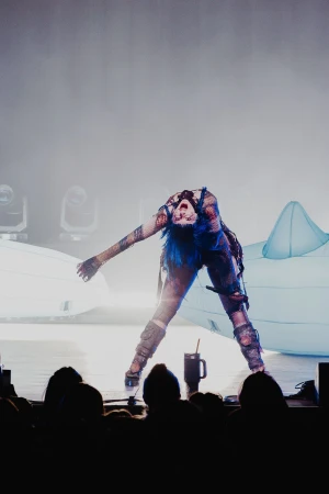 Performer with blue hair and dark makeup bends backward on a stage during a music concert with dark, silhouetted figures in foreground.