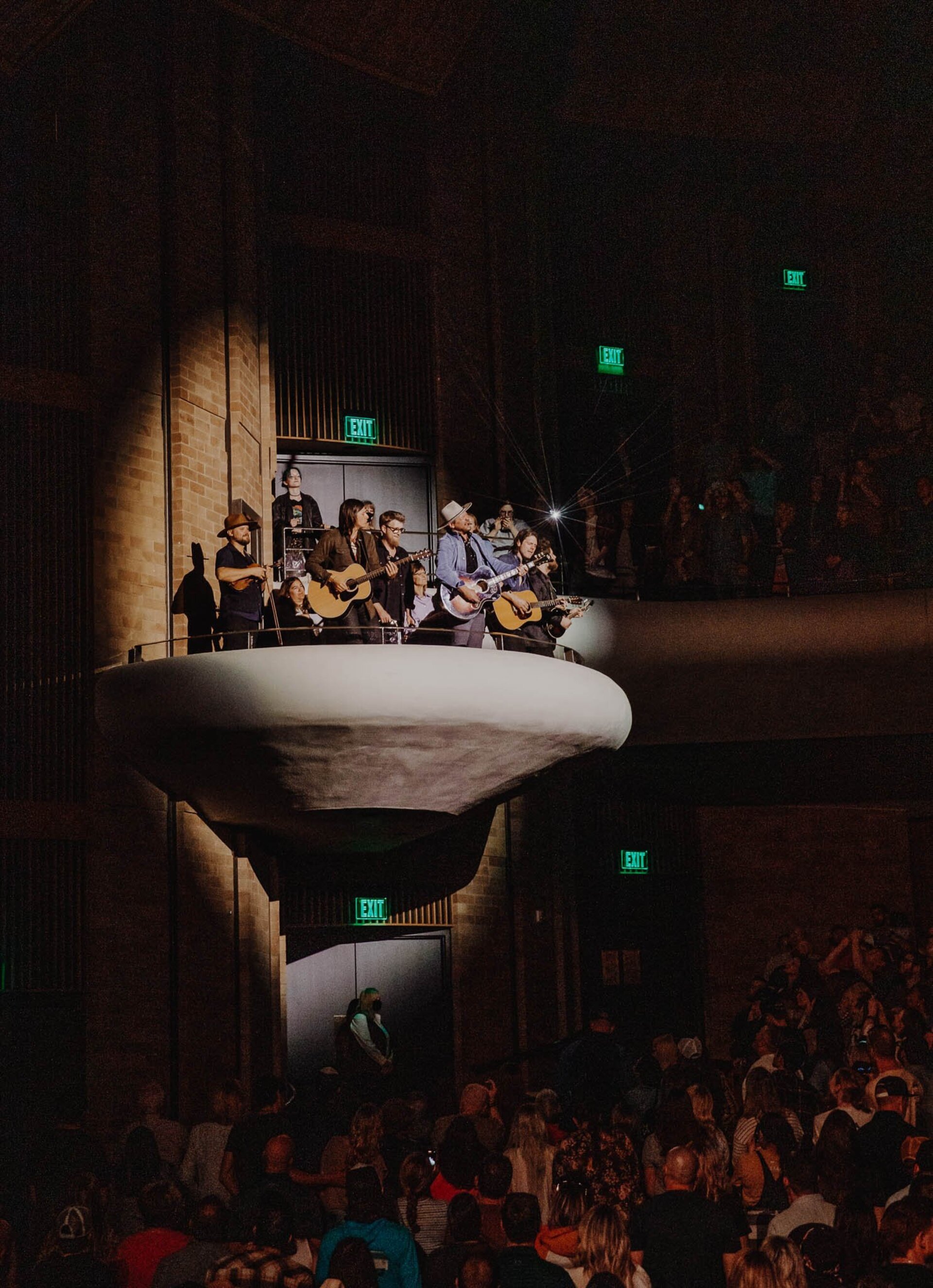 Audience watches a band performing on a suspended platform in a darkened concert hall with exit signs; crowd is looking up at musicians.
