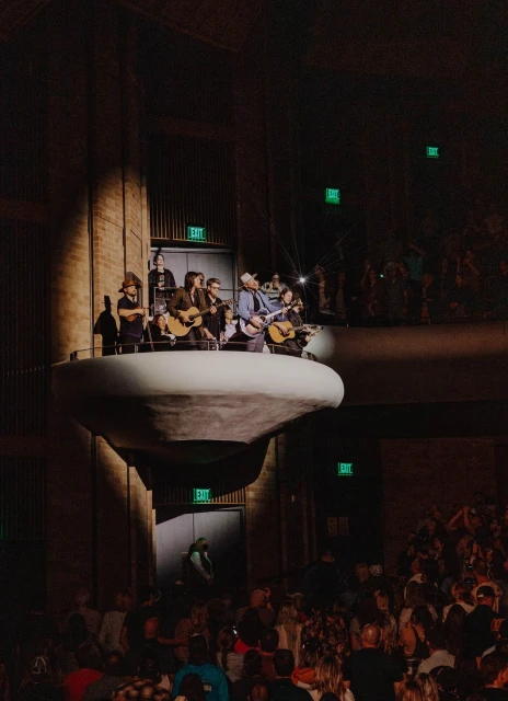 Audience watches a band performing on a suspended platform in a darkened concert hall with exit signs; crowd is looking up at musicians.