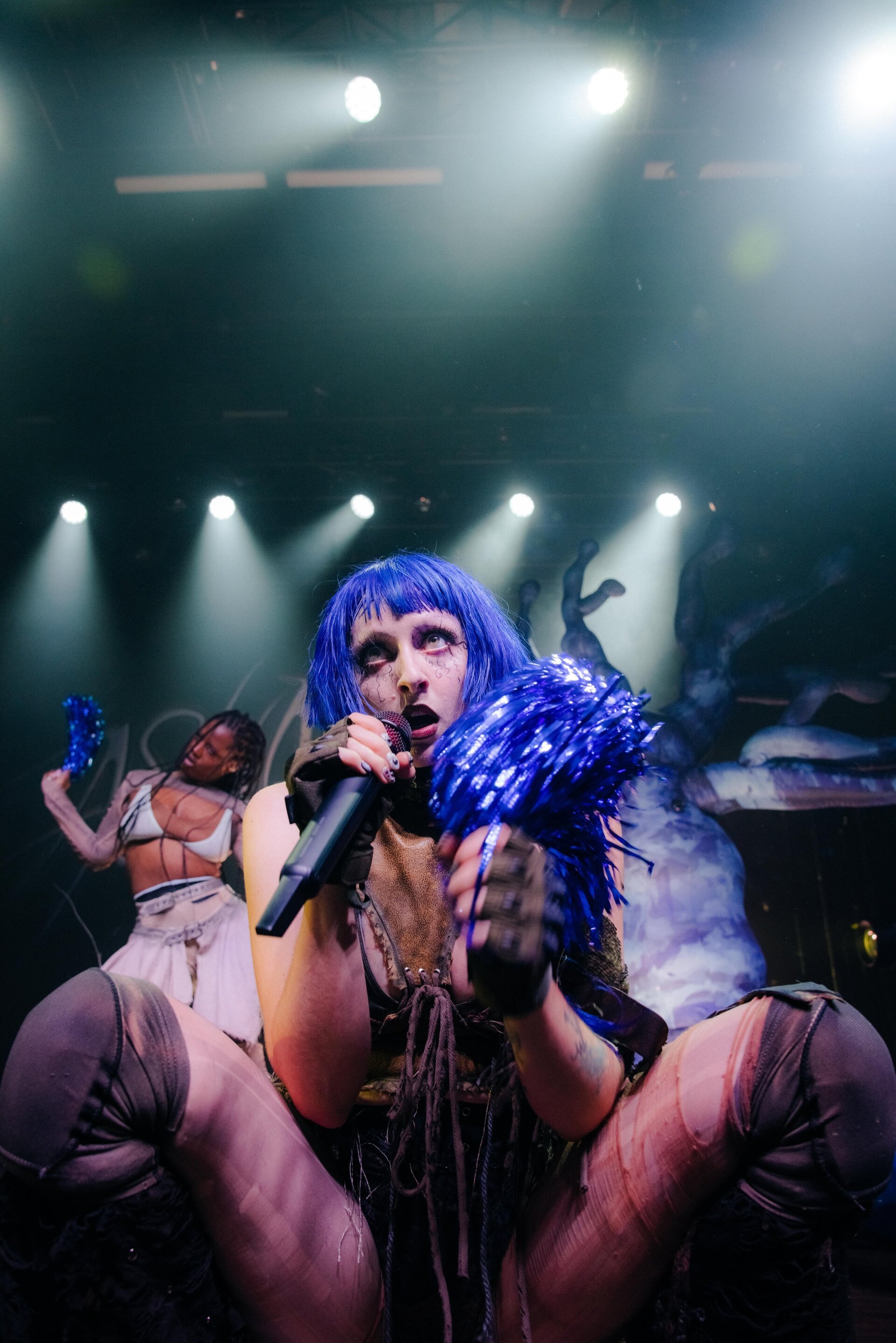 Singer with blue hair performing on stage with a microphone, holding blue pom-poms, and another performer in the background.