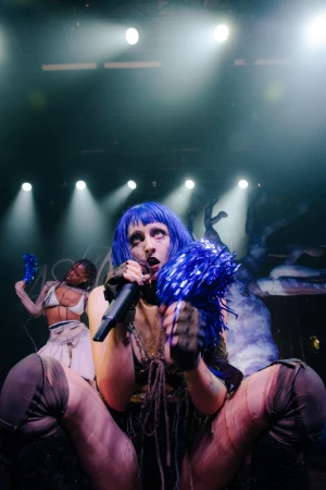 Singer with blue hair performing on stage with a microphone, holding blue pom-poms, and another performer in the background.