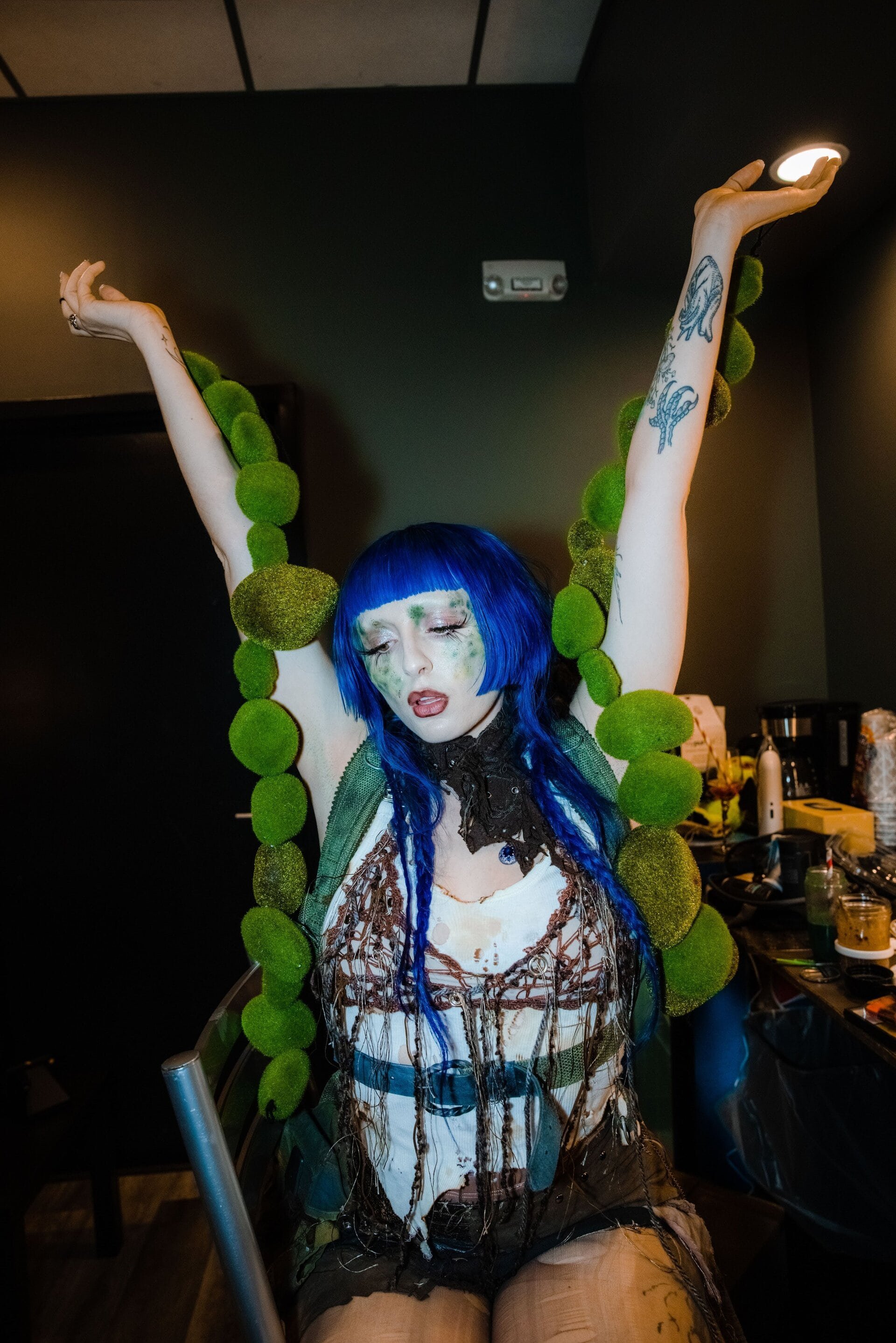 Woman with blue hair and theatrical makeup poses with raised arms, wearing a textured outfit and a large green moss necklace.