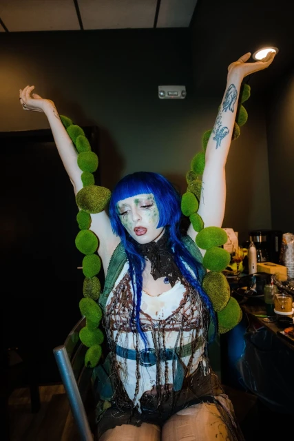 Woman with blue hair and theatrical makeup poses with raised arms, wearing a textured outfit and a large green moss necklace.