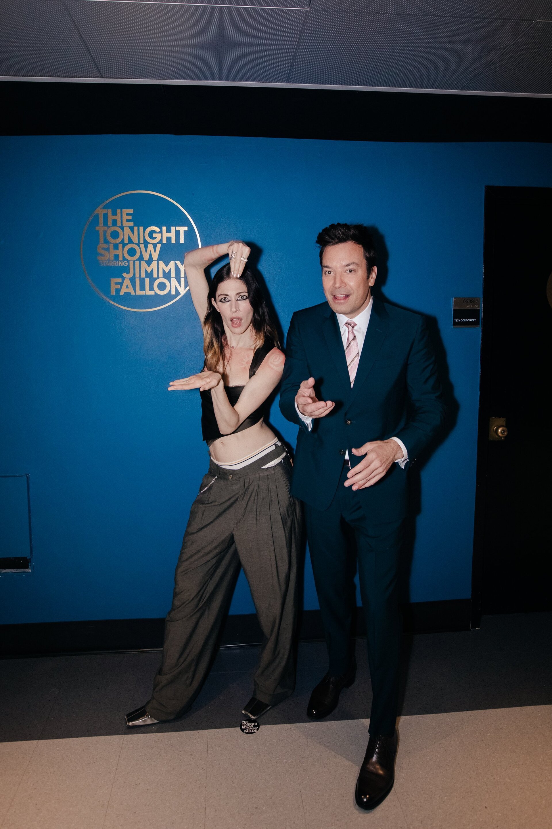 Two people, a woman and Jimmy Fallon, pose in front of a blue wall featuring 'The Tonight Show Starring Jimmy Fallon' logo.