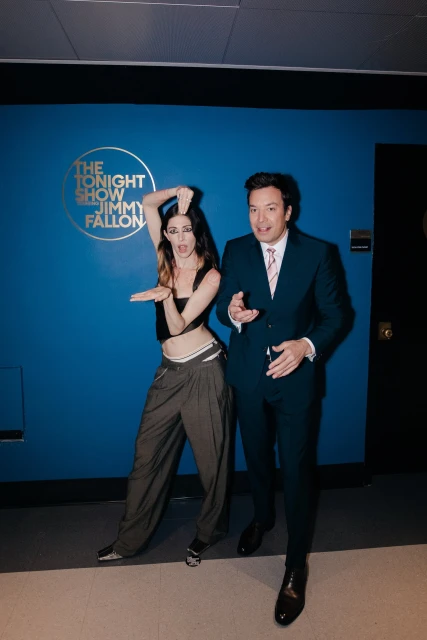 Two people, a woman and Jimmy Fallon, pose in front of a blue wall featuring 'The Tonight Show Starring Jimmy Fallon' logo.