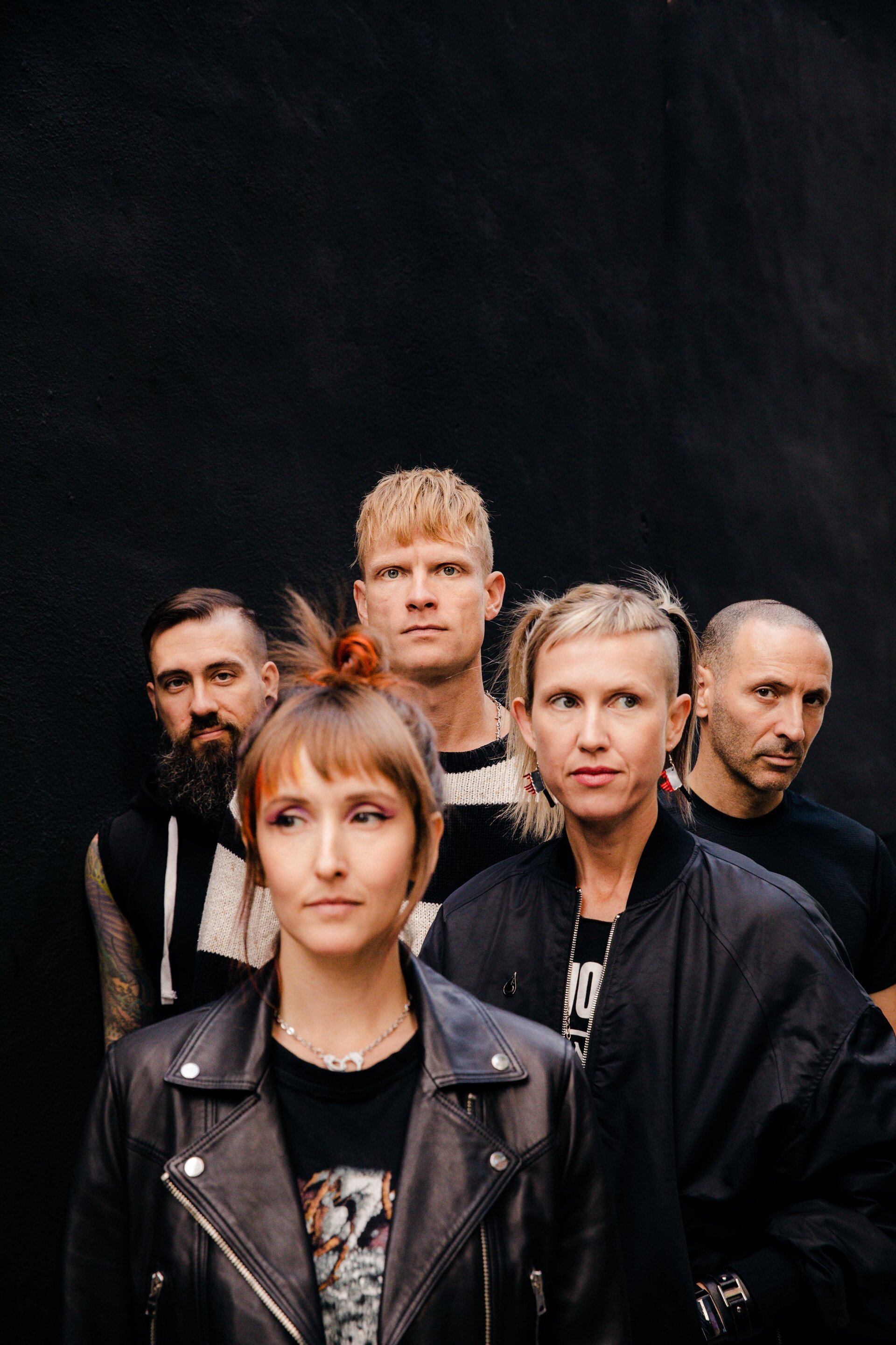 Five adults stand shoulder-to-shoulder indoors against a dark background, wearing casual clothing and showcasing various hairstyles.
