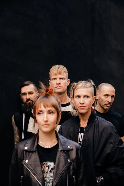 Five adults stand shoulder-to-shoulder indoors against a dark background, wearing casual clothing and showcasing various hairstyles.