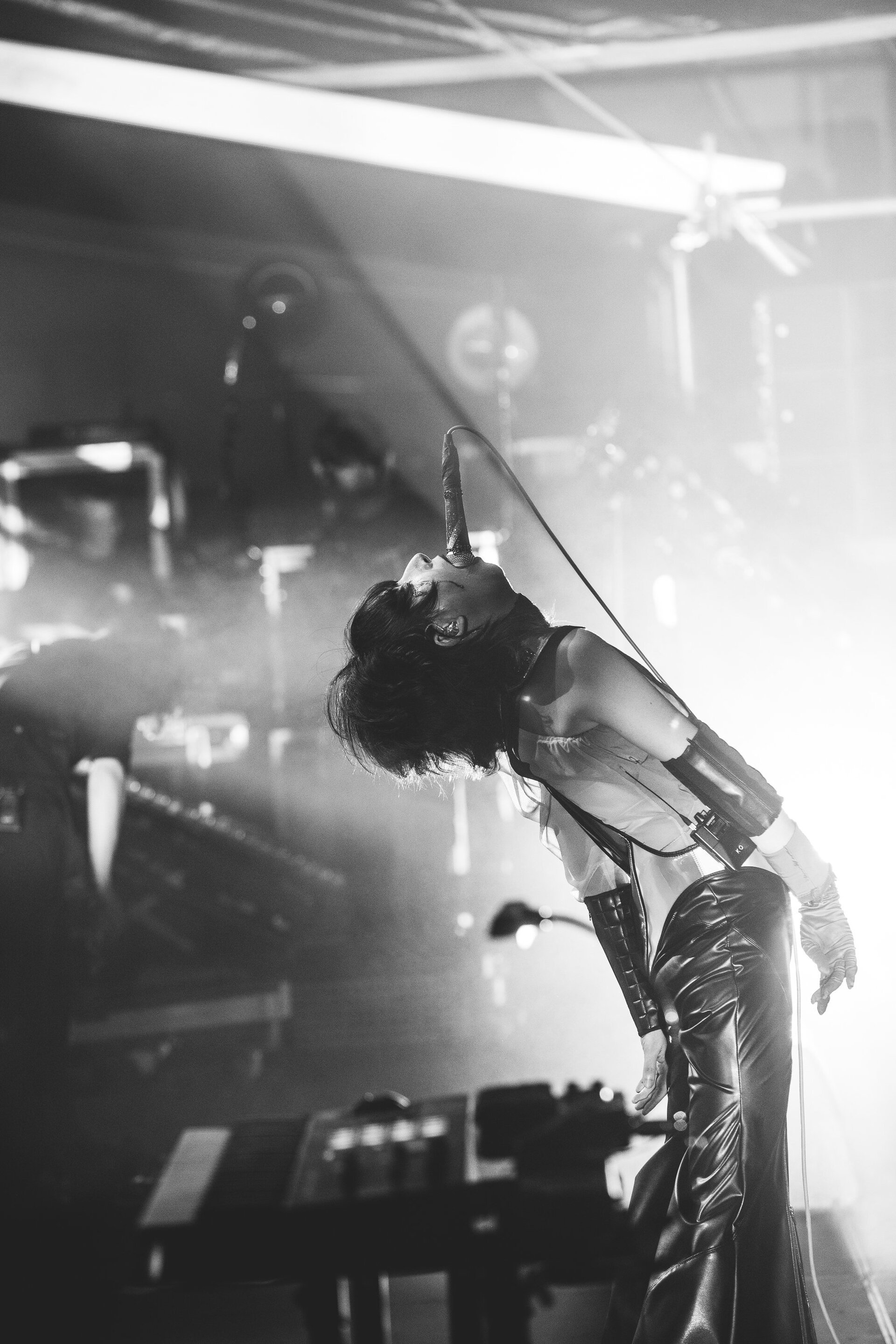 Monochrome stage shot of a performer with dark hair singing into a microphone, wearing a corset and shiny pants in dramatic lighting.