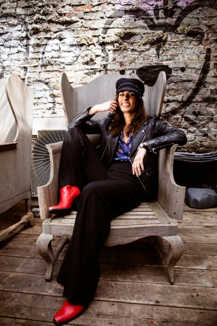 Stylish woman in leather jacket, hat, and red boots poses on a wooden chair against a graffiti brick wall in an urban setting.