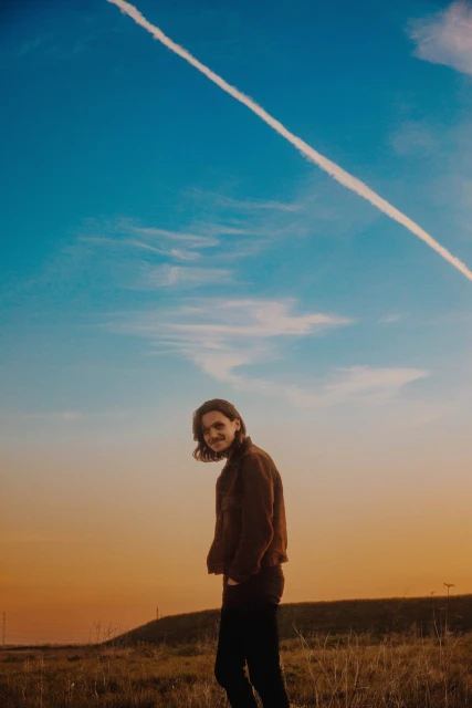 Outdoor portrait of a smiling person in a jacket, standing in a field under a blue sky with contrails at sunset or sunrise.
