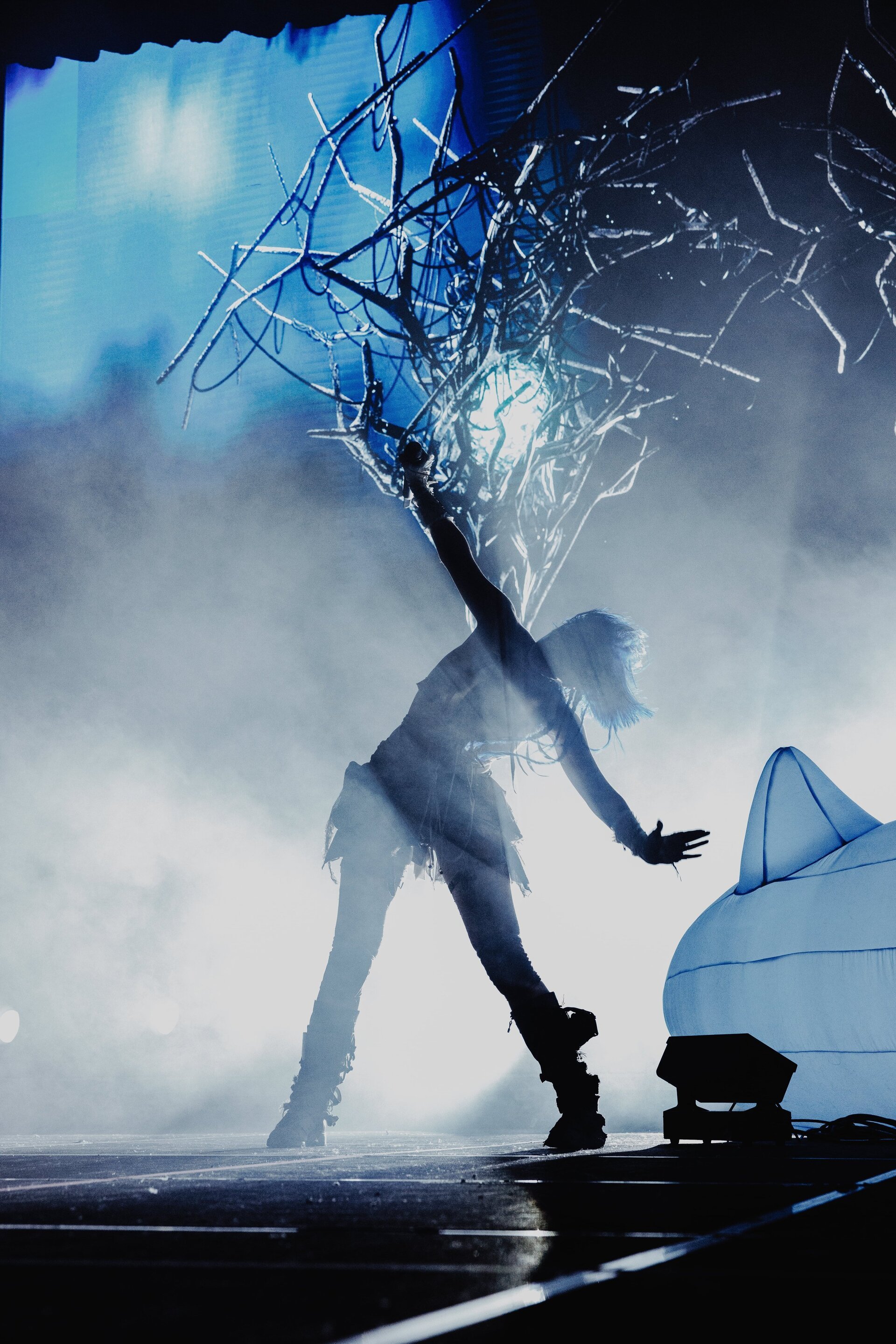 Silhouette of a woman on stage with raised arm, performing with a branch-like prop, under stage lights and a hazy atmosphere.